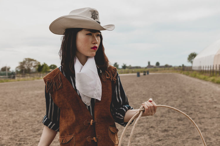 Cowgirl holding rope while standing at ranch against sky