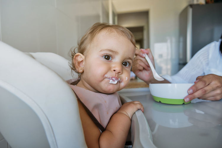 Little girl sitting on a high chair with a bib eating from the spoon with cropped unrecognizable mother feeding her