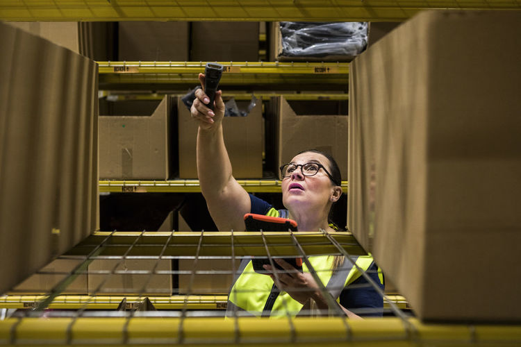 Mature female worker holding digital tablet and bar code reader against rack at distribution warehouse