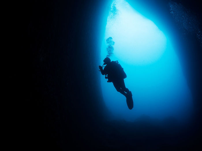 Full body of diver in wetsuit with flashlight in hand swimming underwater of deep sea with clear water near stony formations