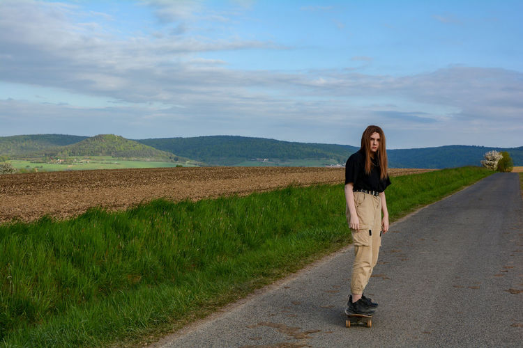 Young girl rides a skateboard on a country road in green nature, with blue sky and many copy space