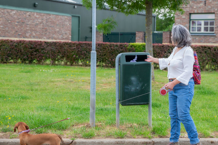 Mature woman in casual clothing depositing her dog's waste in a public rubbish bin on the street