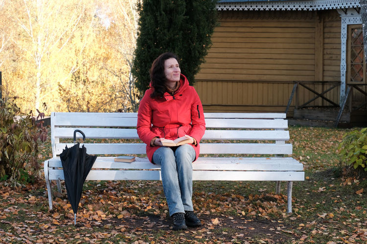 A woman in a red jacket is sitting on a white bench in the park and reading a paper book.
