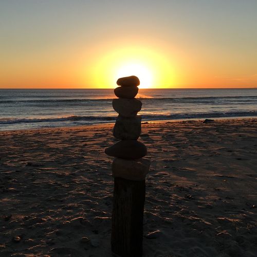 Back lit stones stack on wooden post at beach against sky during sunset