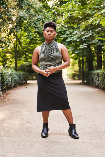 Full body of confident filipino non binary guy wearing top with black skirt with boots standing in pathway of park looking at camera