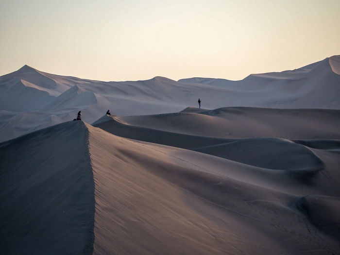 Mid distance view of people standing on sand dunes against clear sky during sunset