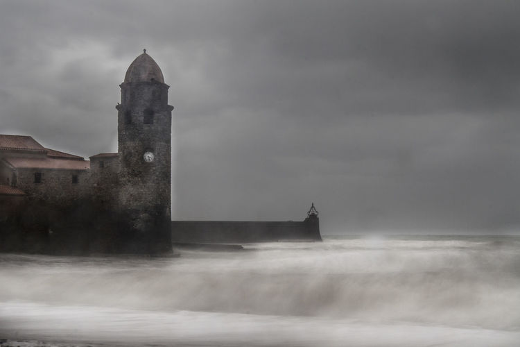Clock tower of eglise de notre dame des anges church by sea against cloudy sky