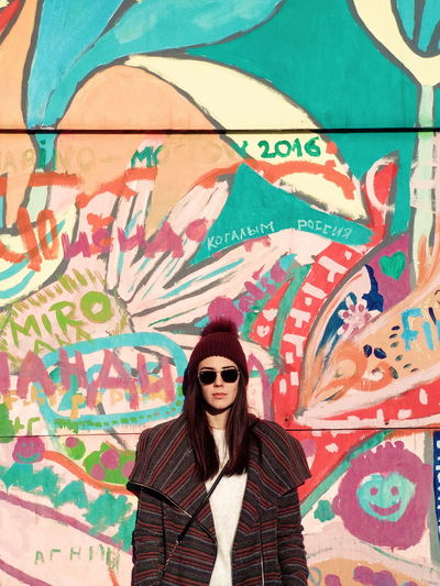 Portrait of young woman wearing sunglasses standing against graffiti wall