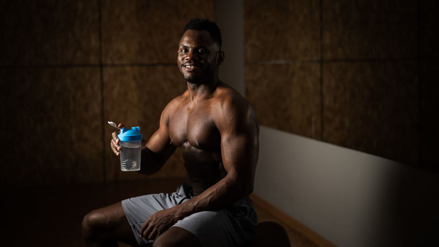 Shirtless man with water bottle sitting against wall