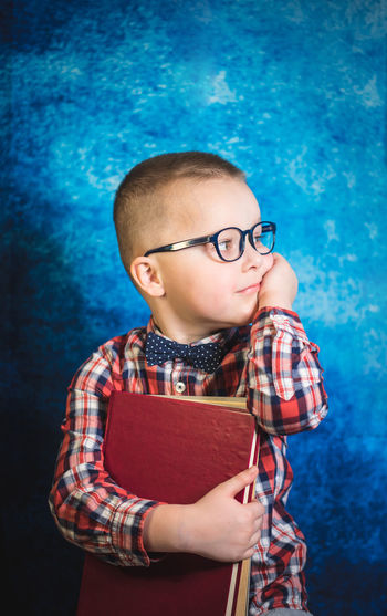 Cute boy wearing eyeglasses and holding book standing against blue wall