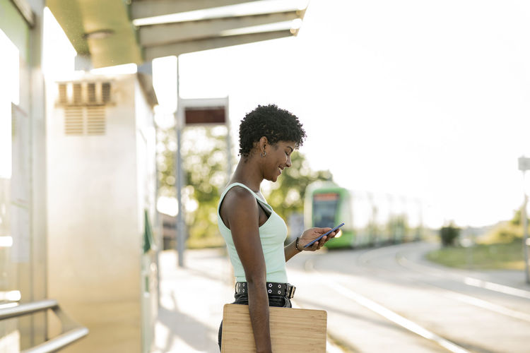 Smiling young woman with laptop using smart phone while standing at tram station