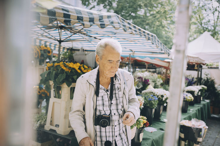 Senior tourist wearing camera shopping at flower market in city during vacation