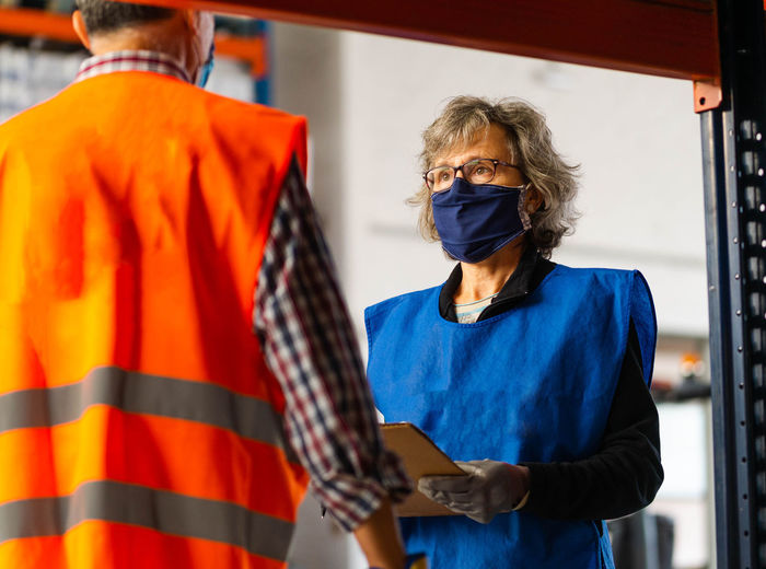 Serious storehouse specialists wearing uniforms and protective masks standing near shelves in modern warehouse and discussing work process