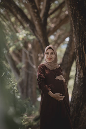 Portrait of pregnant woman standing by tree trunk in forest