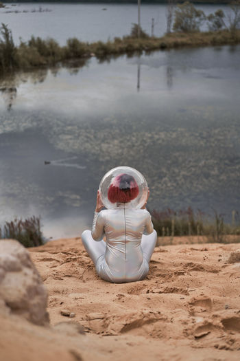 Back view of unrecognizable futuristic young red haired female in silver space suit and glass helmet sitting on sand near flooding river