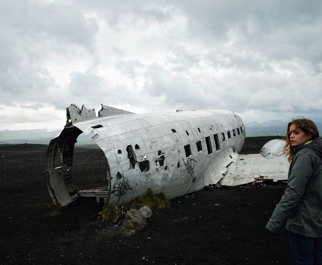 Young woman standing by abandoned airplane on field against cloudy sky