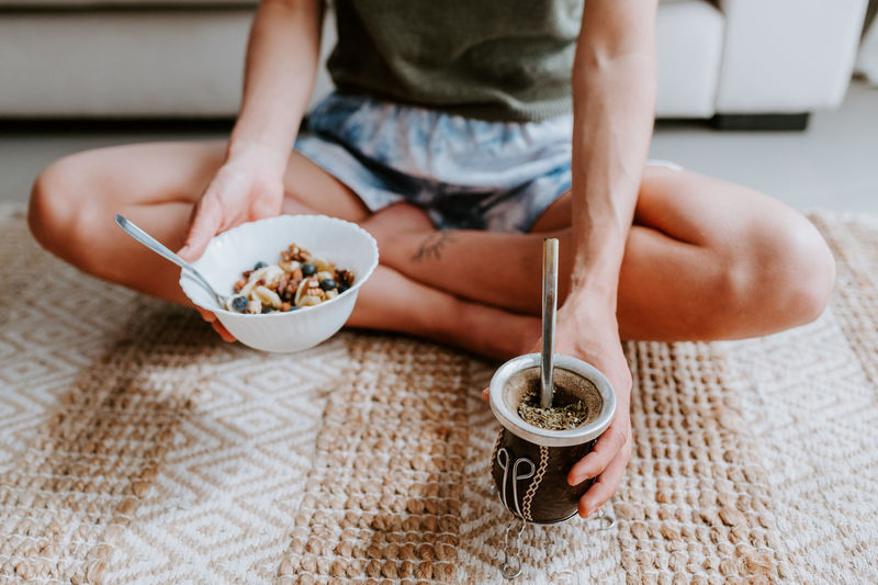 Cropped unrecognizable female with tasty mate tea drink and bowl of healthy granola sitting on floor at home and having nourishing breakfast