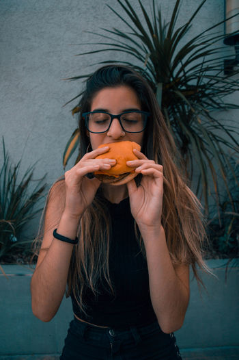 Young woman eating burger while standing against wall