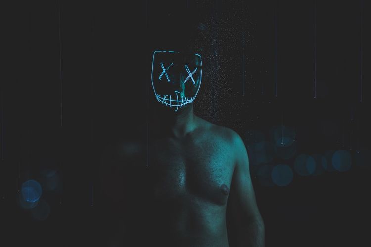 Shirtless man wearing spooky mask while standing in darkroom