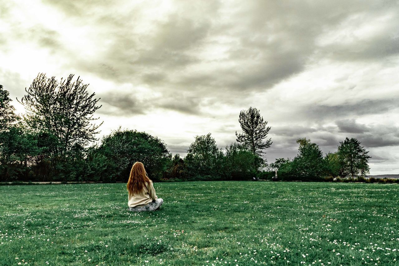 REAR VIEW OF WOMAN SITTING BY GRASS AGAINST SKY
