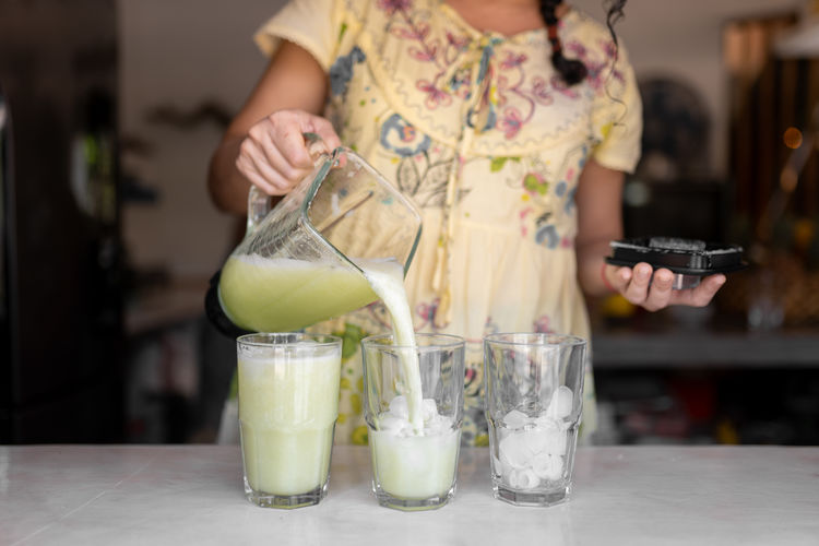 Crop anonymous female in dress pouring healthy cucumber smoothies from blender jar into glasses placed on table in light kitchen