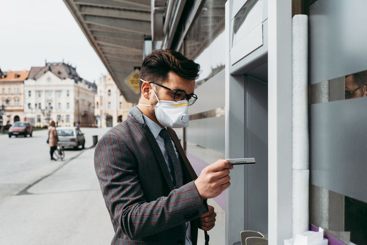 Business man with protective face mask using street atm machine.