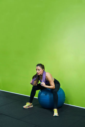 Tired sweaty hispanic female athlete sitting with towel on neck on fit ball after active workout in gym