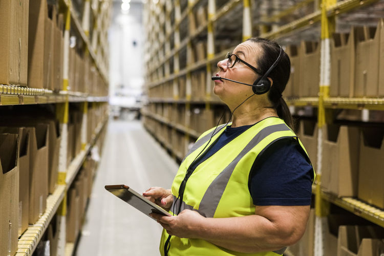 Mature female worker with digital tablet looking up while talking on headset at distribution warehouse