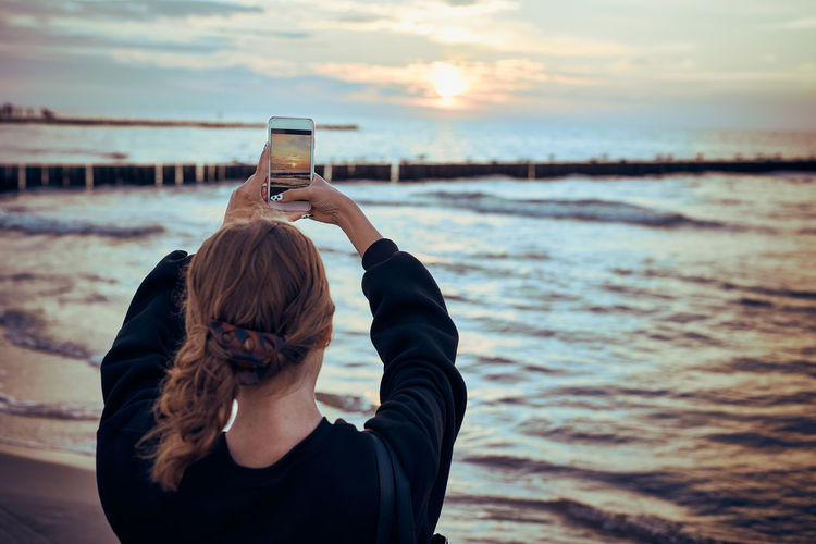 Young woman taking photos of sunset over sea using smartphone during summer trip