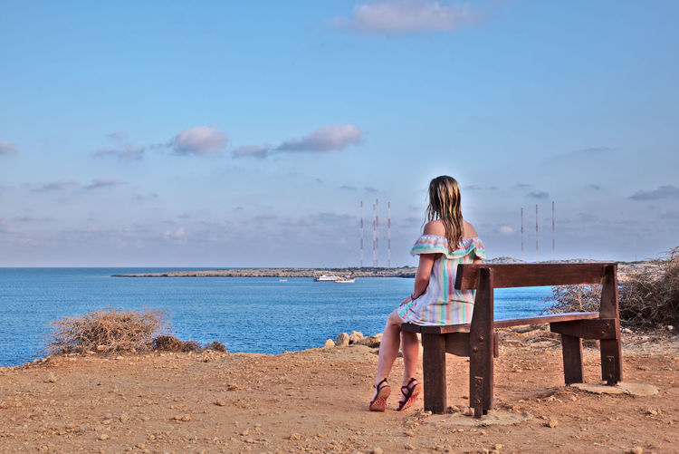 Rear view of woman sitting on bench at beach against sky