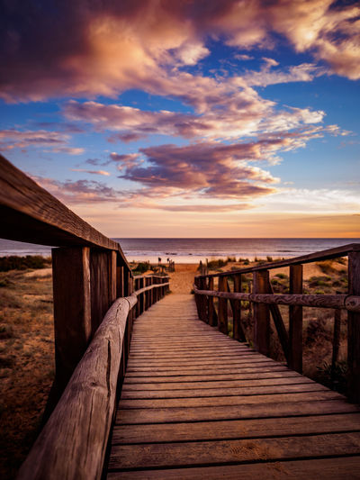 Wooden walkway leading towards sea against sky during sunset