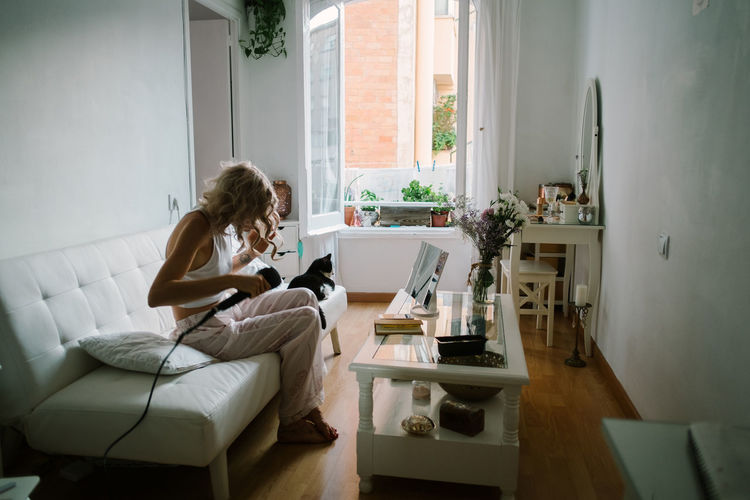 Side view of unrecognizable female in domestic wear sitting on sofa at home and curling hair with iron