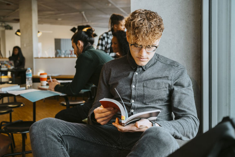 Young blond man reading book while sitting in university cafeteria