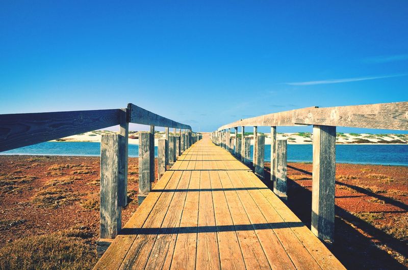 Wooden footbridge leading towards sea against clear blue sky