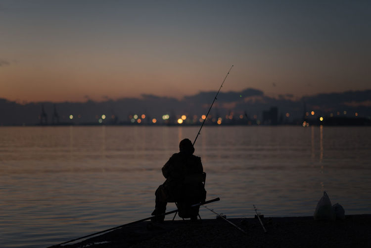 Rear view of silhouette man fishing by lake against sky during sunset