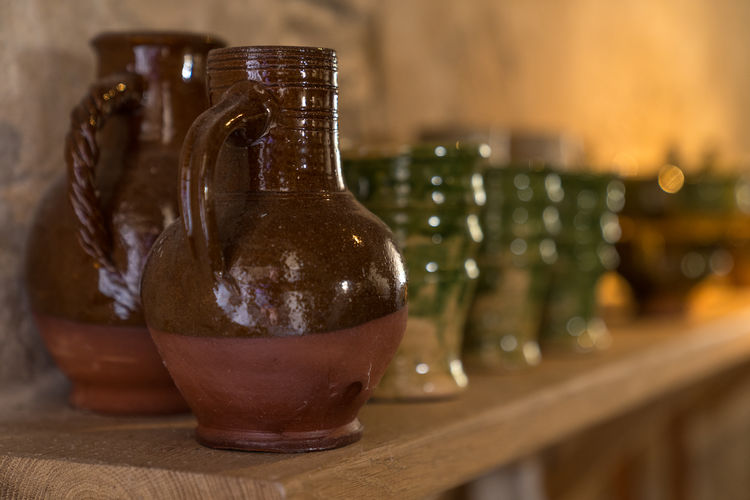 Many medieval earthenware jugs and cups in brown green glazing on wooden shelf near stone wall