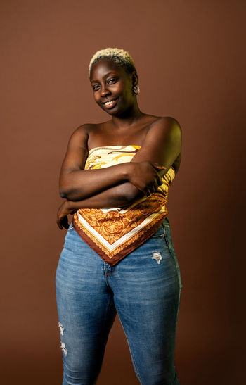 Happy african american female model wearing silk scarf top with arms folded on brown background in studio and looking at camera