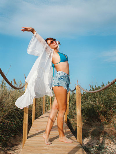 Young woman in white cover up and jeans shorts on a walkway over the dunes