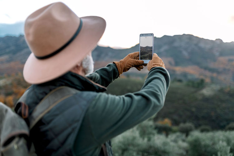 Back view of unrecognizable senior male explorer taking photo of mountainous landscape while using smartphone and enjoying vacation in caceres