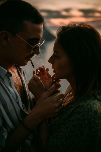 Man lighting cigarette for girlfriend while chilling on seashore at sunset