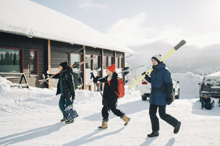 Full length of friends walking with skis at tourist resort during sunny day