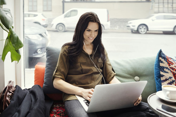 Smiling businesswoman using laptop while sitting on sofa against window