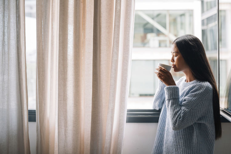 Young woman with cup of coffee standing in front of open window