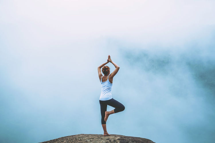 Full length rear view of woman with arms raised doing yoga o rock against sky