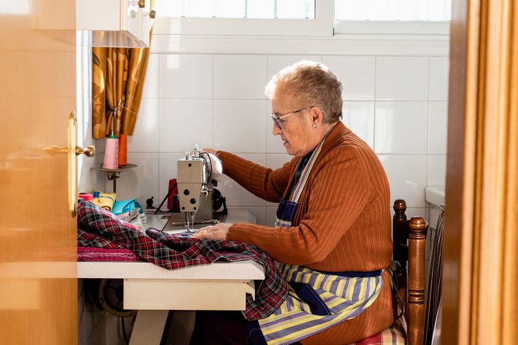 Side view of concentrated senior seamstress sitting at sewing machine and stitching piece of cloth while working in workshop