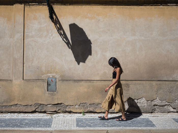 Side view of woman sitting on sidewalk against wall