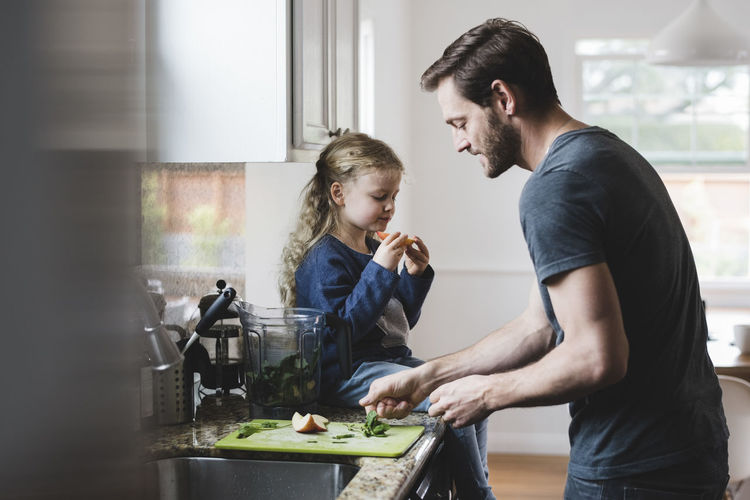 Side view of father cooking food while daughter having apple in kitchen