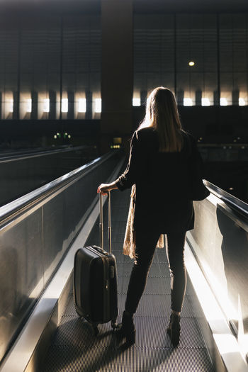 Woman with wheeled luggage moving on escalator at station