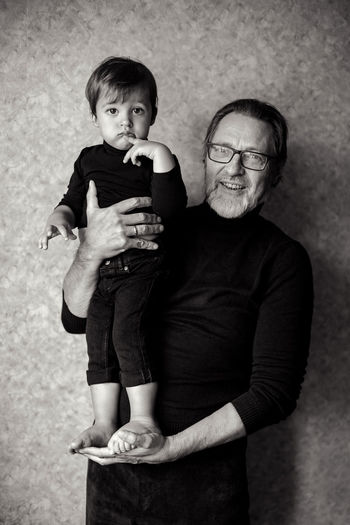 Grandfather with beard and glasses with his grandson sitting on his hands in the studio