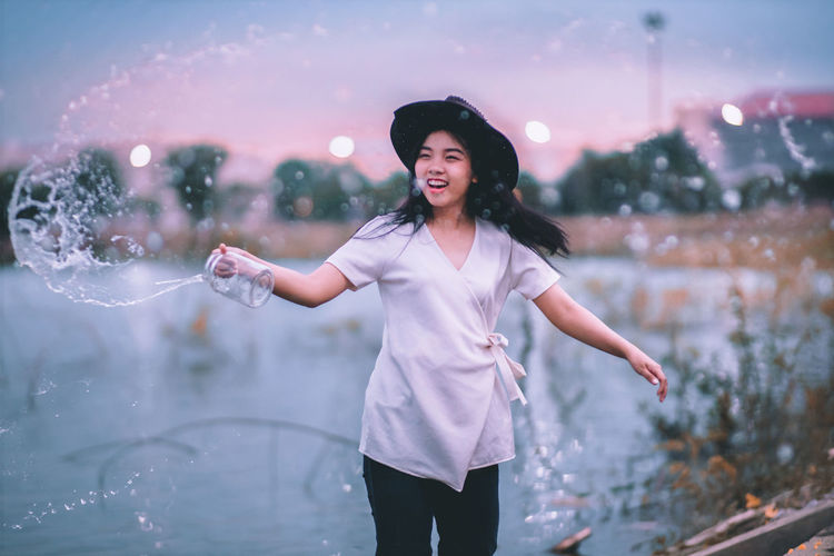 Happy young woman spraying water while standing by lake against sky during sunset